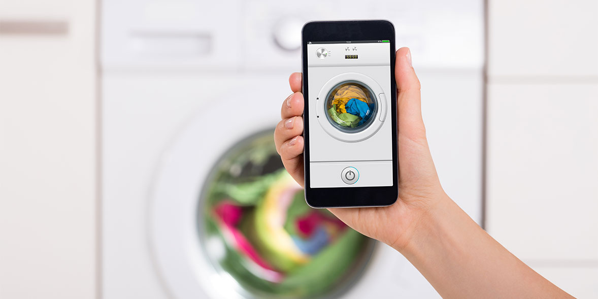 A hand holds a smartphone displaying a washing machine control app, with laundry visible inside the machine.
