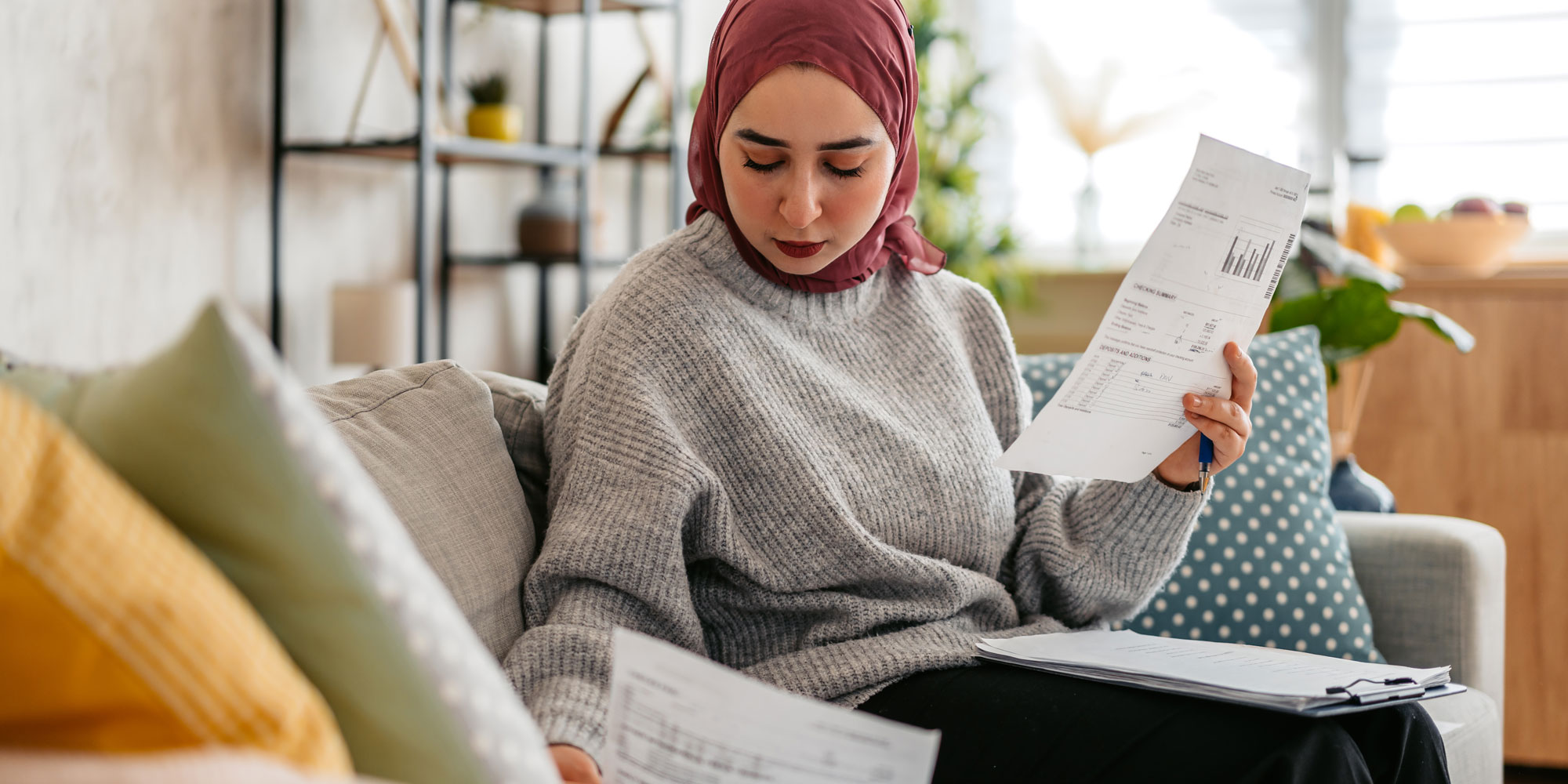 A mother and her child sit on a couch, happily inserting coins into a pink piggy bank together.