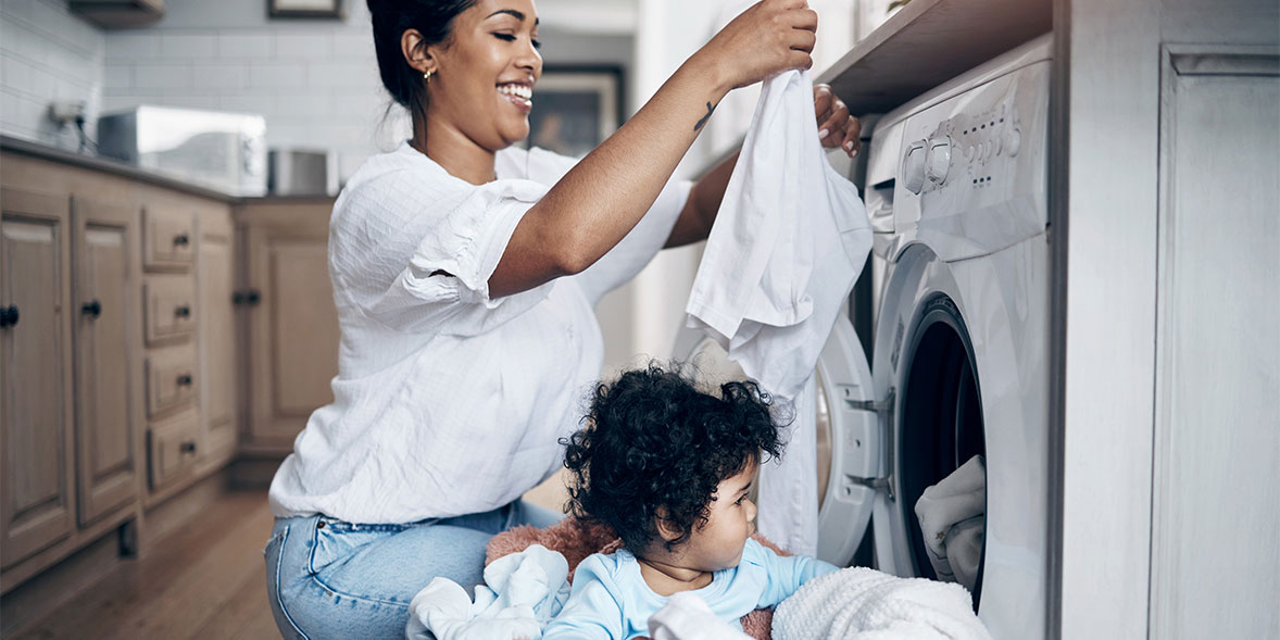 Woman smiling while placing clothes in a washing machine, with a toddler seated nearby surrounded by laundry.