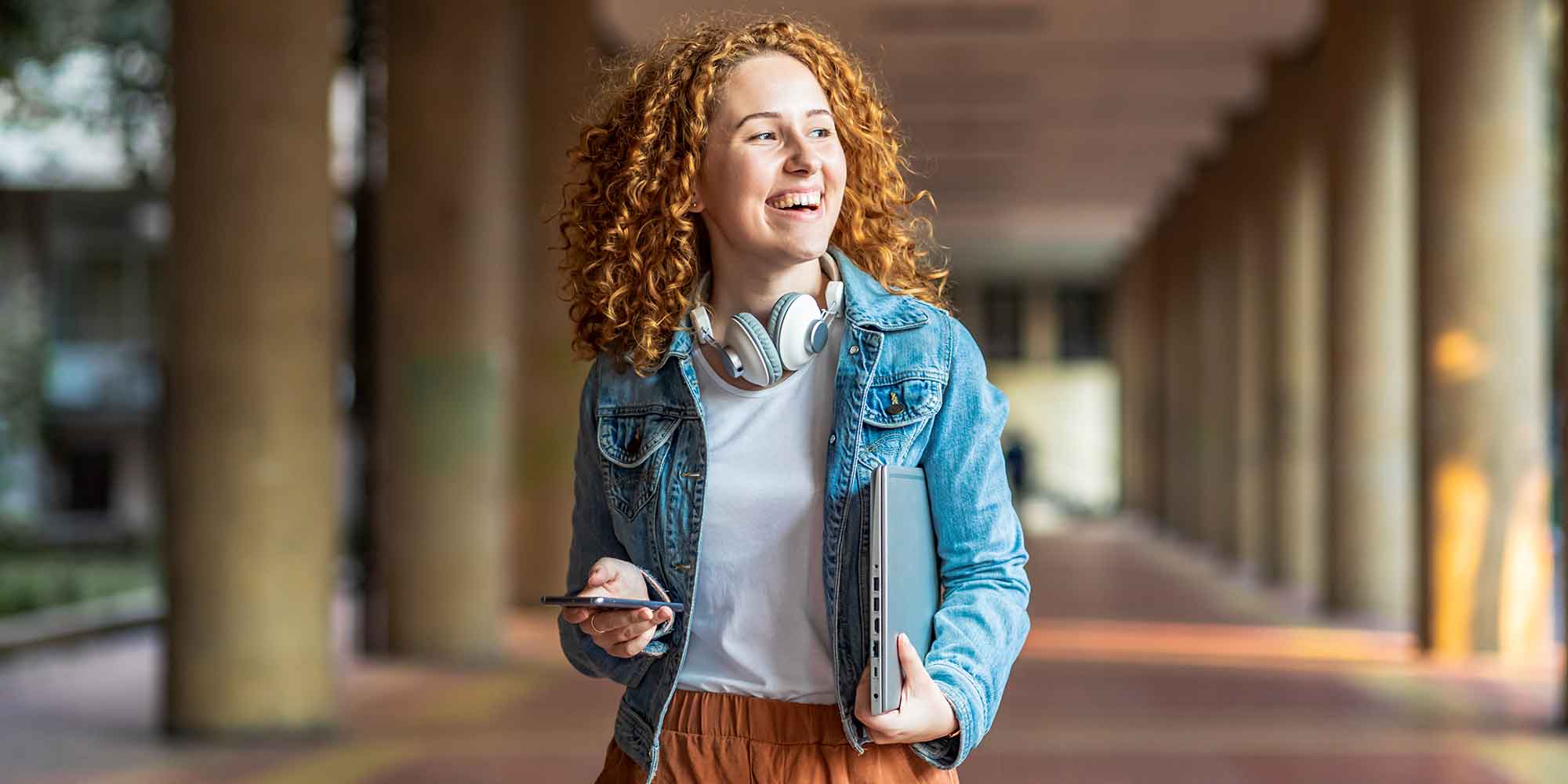 Young man wearing headphones studies at a desk in a library, using a laptop and taking notes in a notebook.