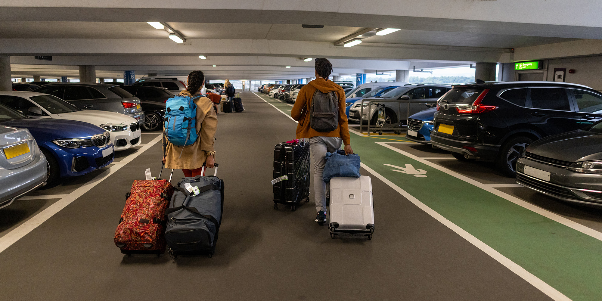 Two travelers walk through a parking garage, pulling luggage, surrounded by parked cars on either side.