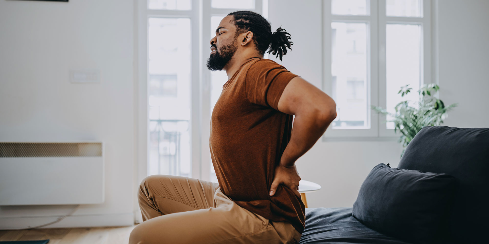 Man sitting on a sofa, leaning forward with a pained expression, holding his lower back, in a bright, airy room.