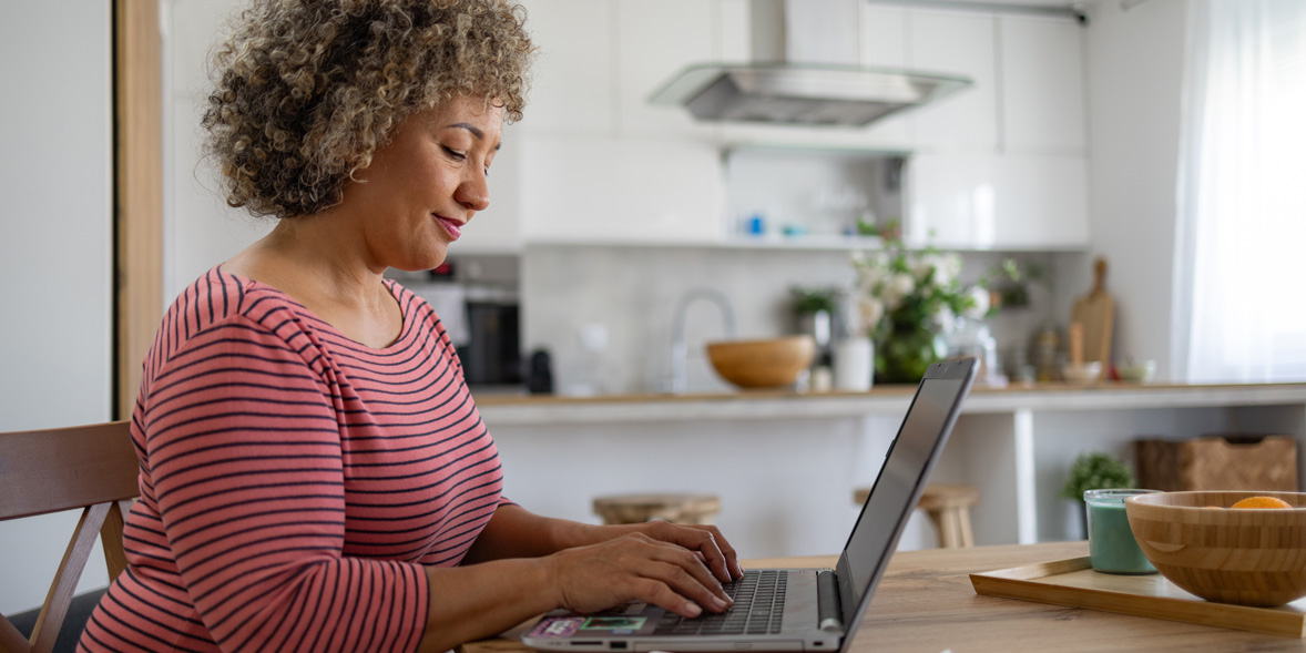 A woman with curly hair types on a laptop at a wooden table, surrounded by a cozy kitchen and plants.