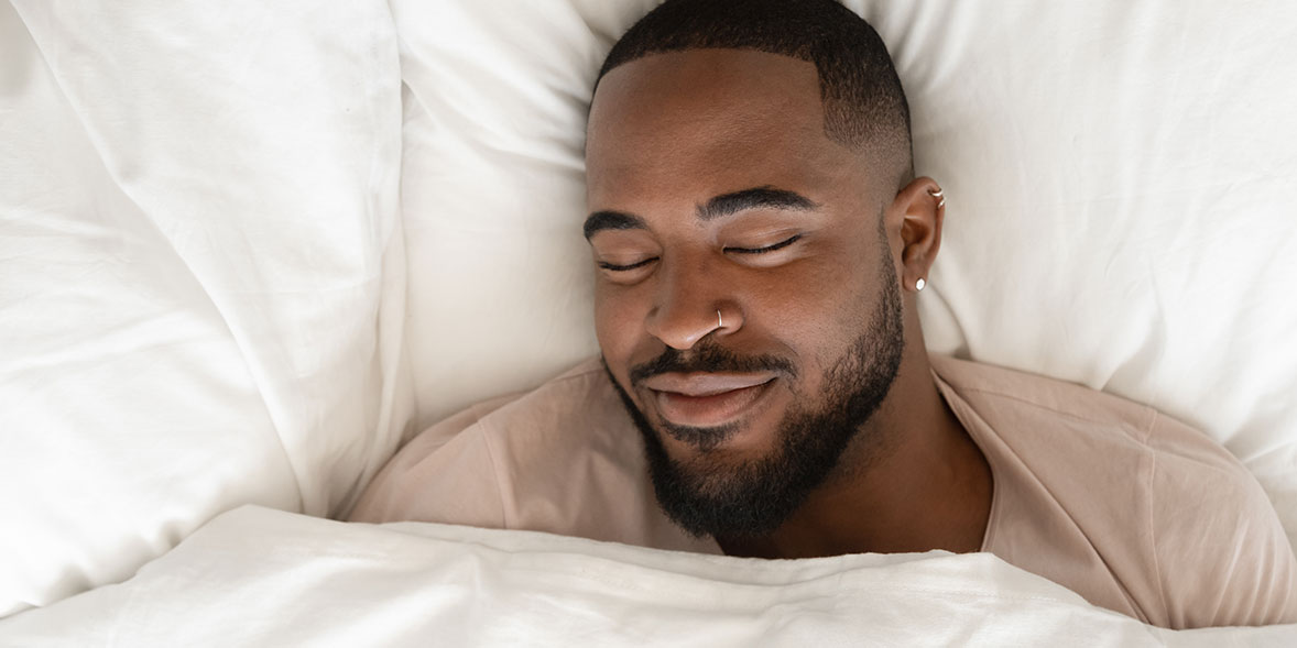 Smiling man with a beard, eyes closed, resting peacefully under white blankets in a cozy bed.