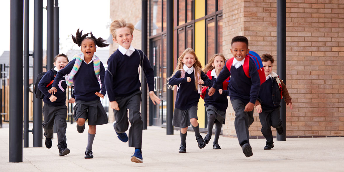 Seven children in school uniforms run excitedly outdoors, smiling, with a modern building in the background.