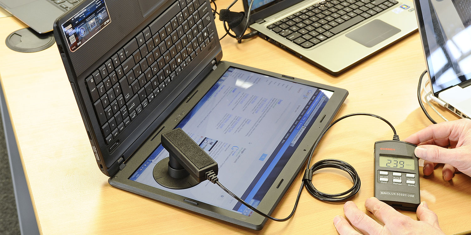 A laptop is positioned upright with a device connected to its screen, measuring brightness while displaying a webpage.