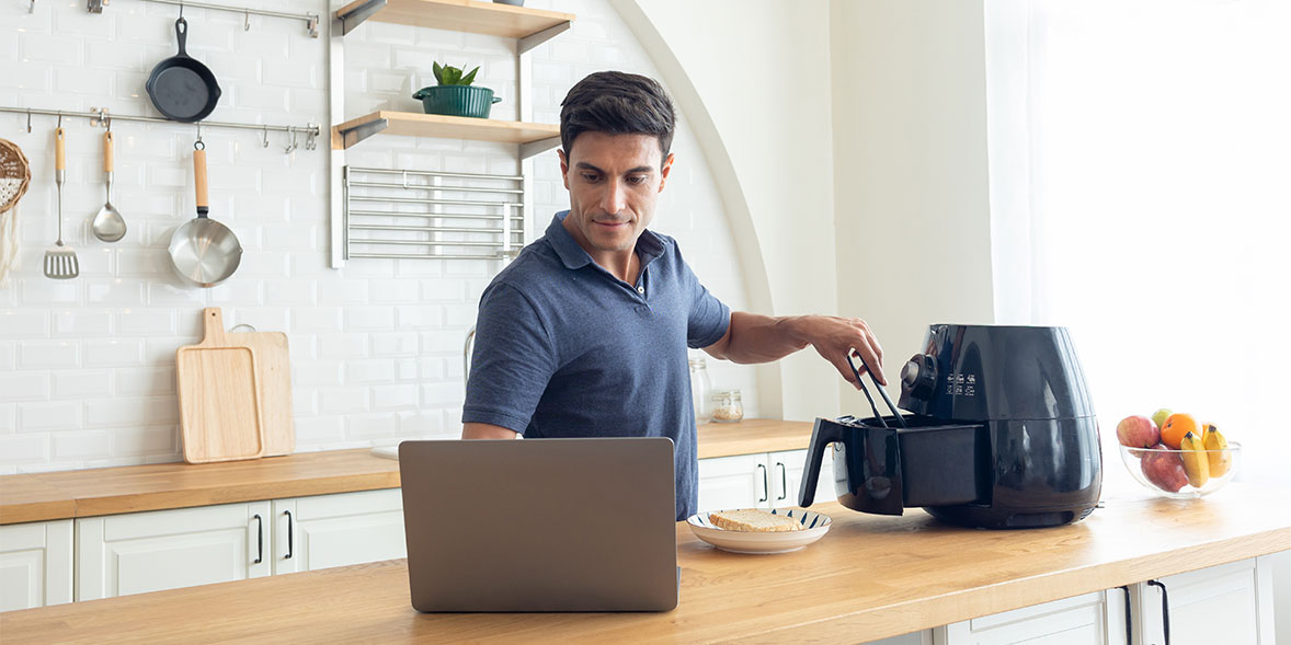 Man cooking in a modern kitchen, using an air fryer while checking a laptop on the countertop.