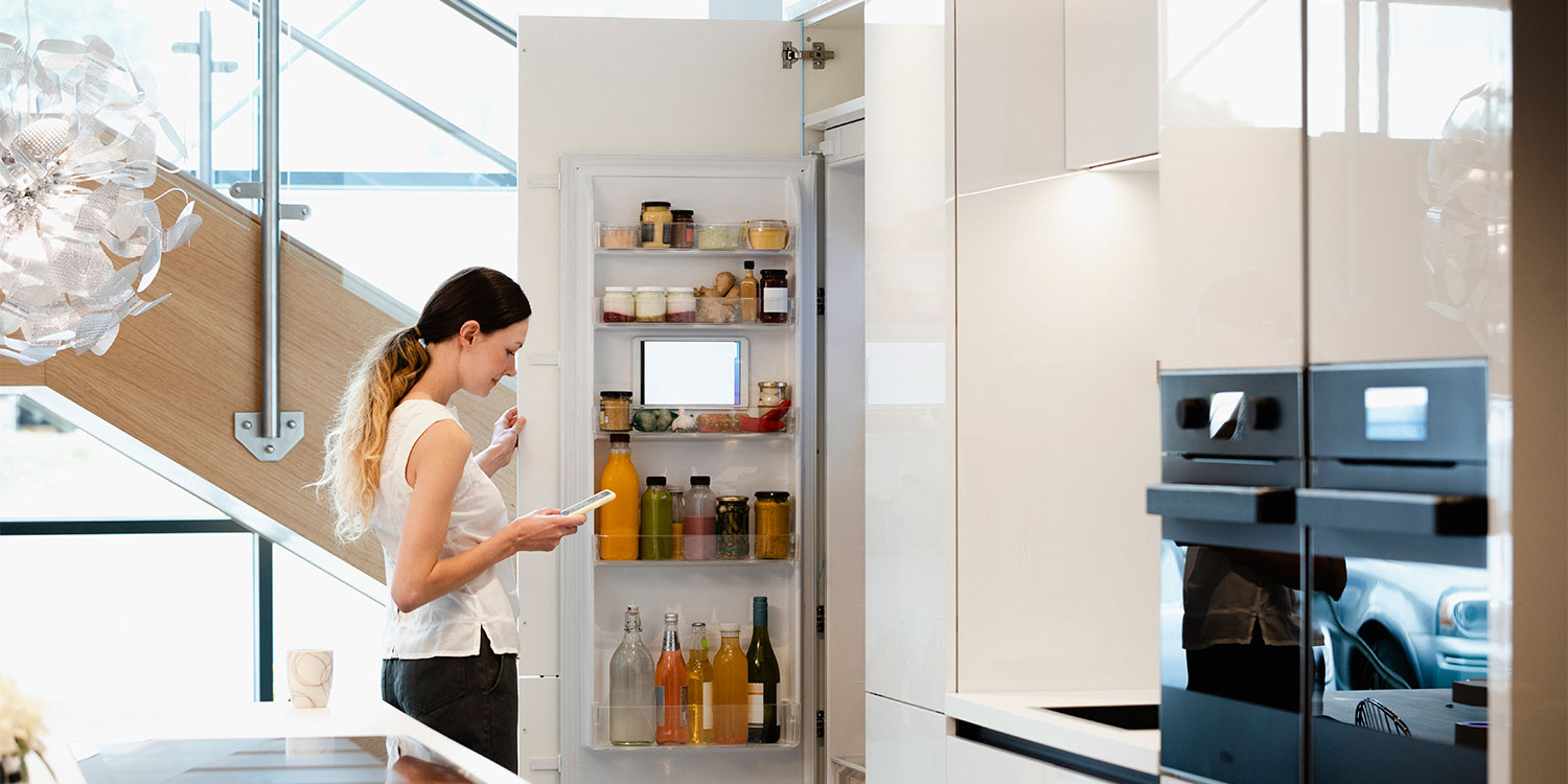 Woman stands in a modern kitchen, looking at her phone while checking an open refrigerator filled with jars and drinks.