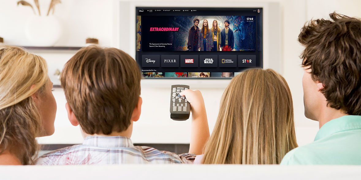 Three people sit on a couch, facing a TV displaying a streaming service menu with various show logos. One holds a remote.