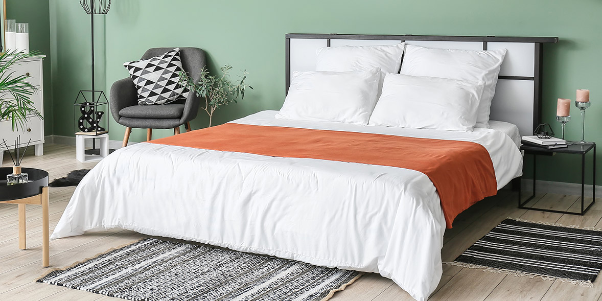 A cozy bedroom featuring a white bed with an orange throw, a patterned rug, a green wall, and a chair with a geometric pillow