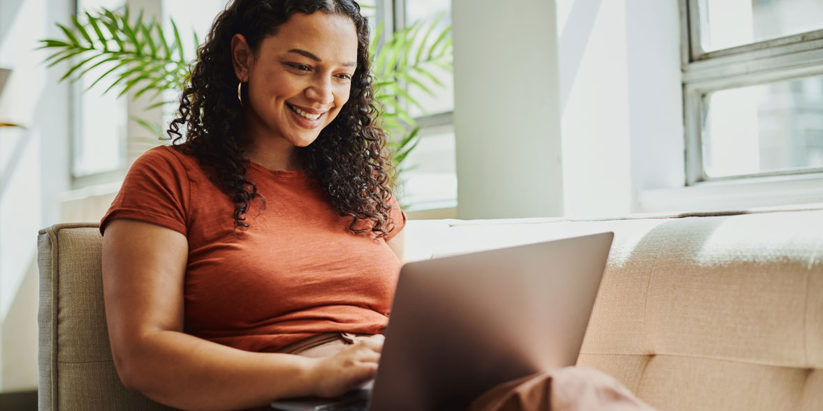 Smiling woman with curly hair using a laptop on a beige couch, surrounded by natural light and greenery.