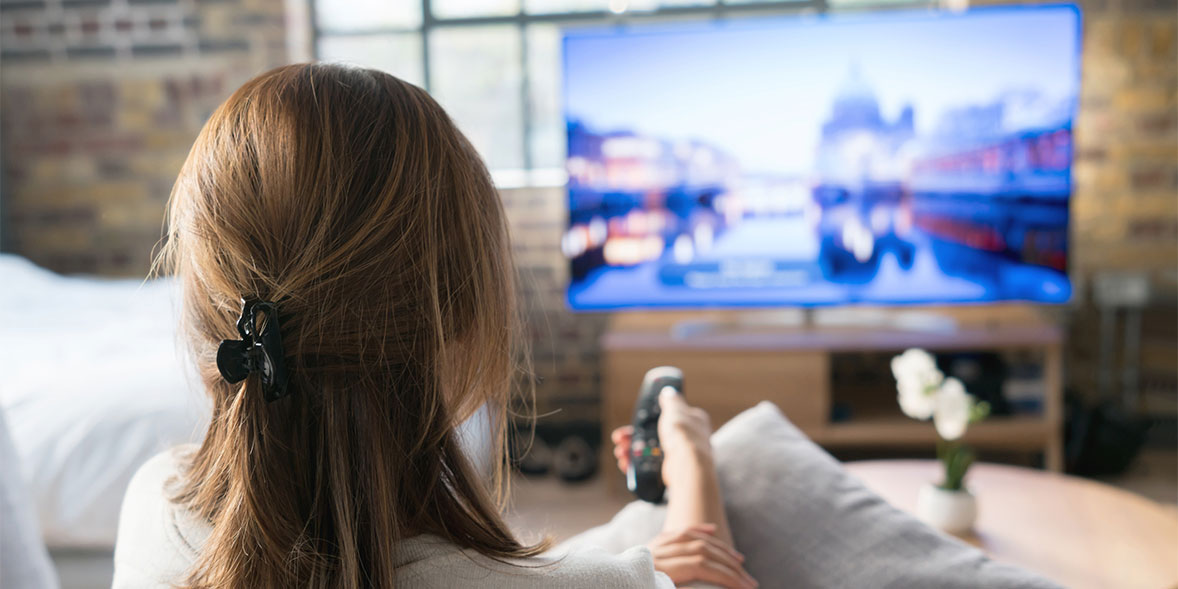 Woman with long hair in a clip sits on a couch, holding a remote, watching a blurred cityscape on a large TV.