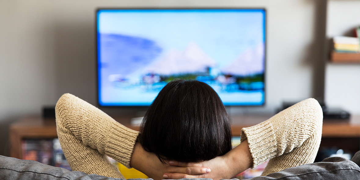 Person with dark hair relaxes on a couch, hands behind their head, watching a blurred scenic landscape on a television.