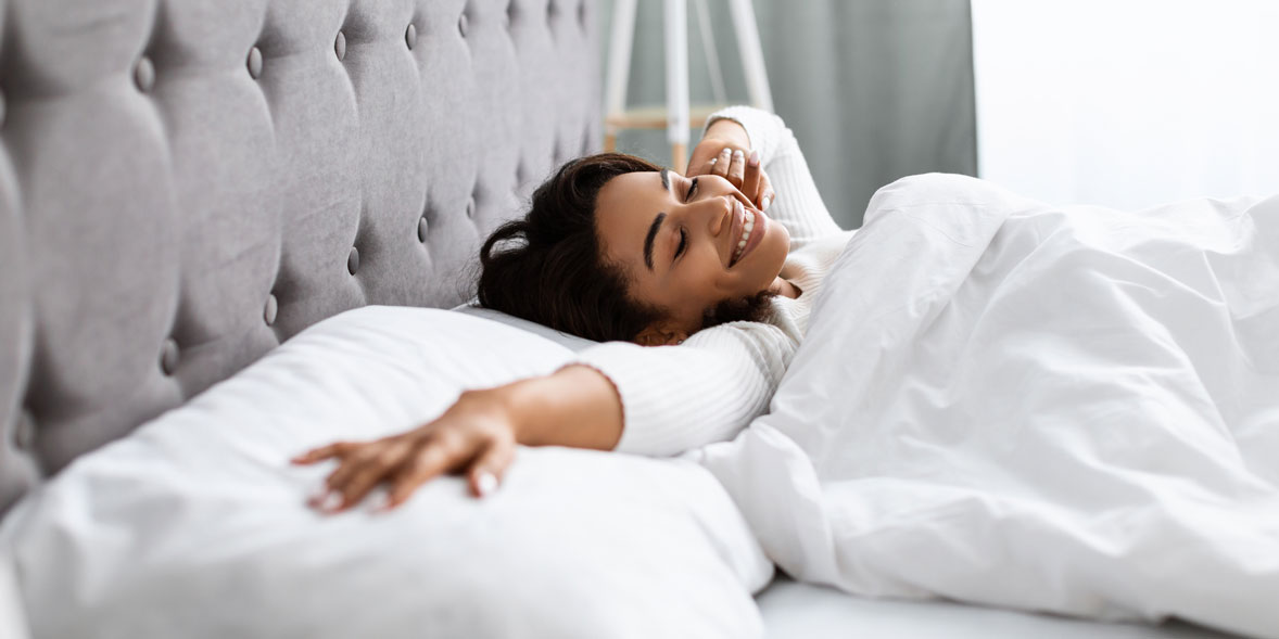 Smiling woman stretching in bed, covered with white sheets, with a cozy grey headboard in a softly lit room.