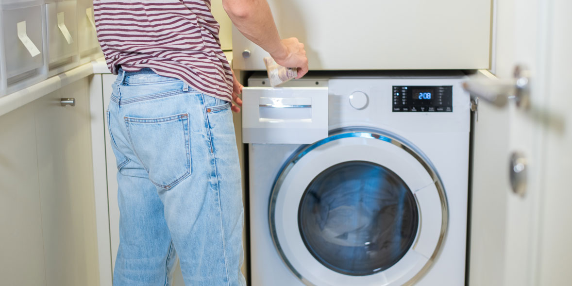 A person in a striped shirt inserts laundry detergent into a front-loading washing machine with a digital display.