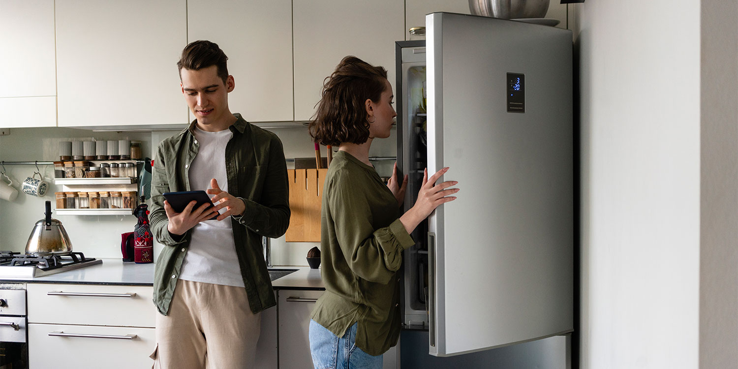 Man using a tablet stands beside a woman looking into a fridge freezer in a modern kitchen with light colors.