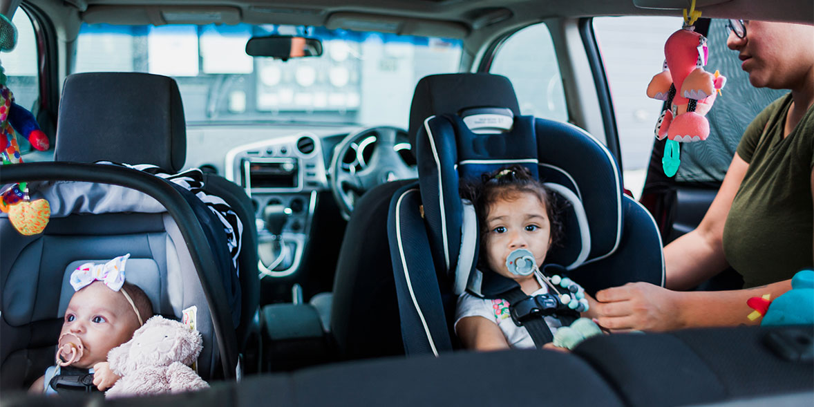 A toddler in a car seat with a pacifier smiles, while an infant sleeps in a nearby car seat.