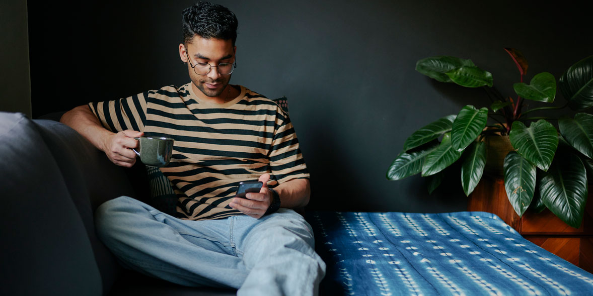 Man in striped shirt sits on a couch, holding a cup and looking at his phone, with greenery in the background.