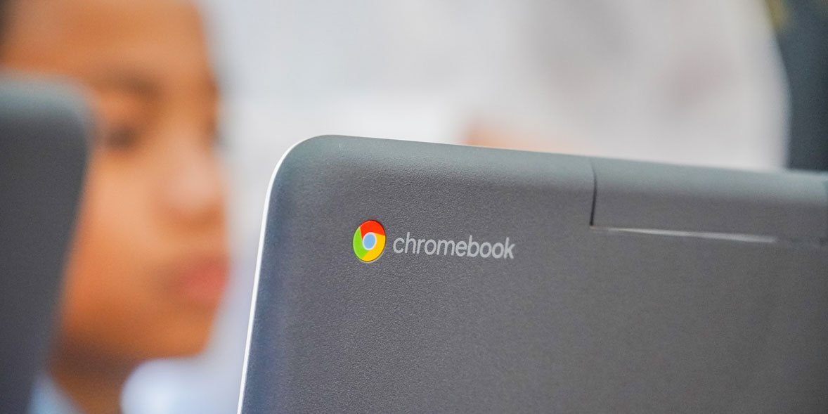 Close-up of a Chromebook with the logo visible; a blurred child’s face in the background, suggesting a learning environment.