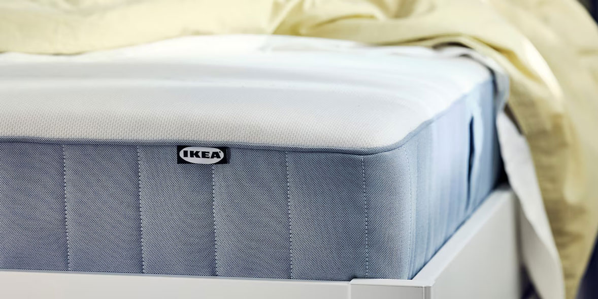 Mattress on a wooden bed frame, partially covered by a light yellow blanket. An IKEA label is visible on the side.
