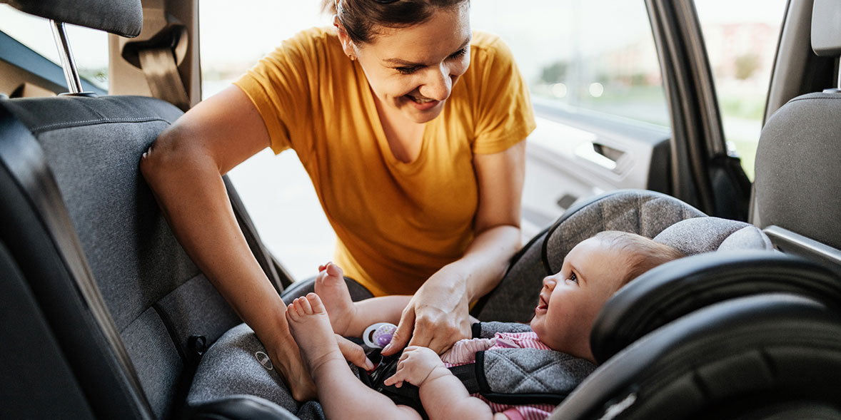 A woman in a yellow shirt smiles at a happy baby in a car seat, adjusting the harness in a vehicle.