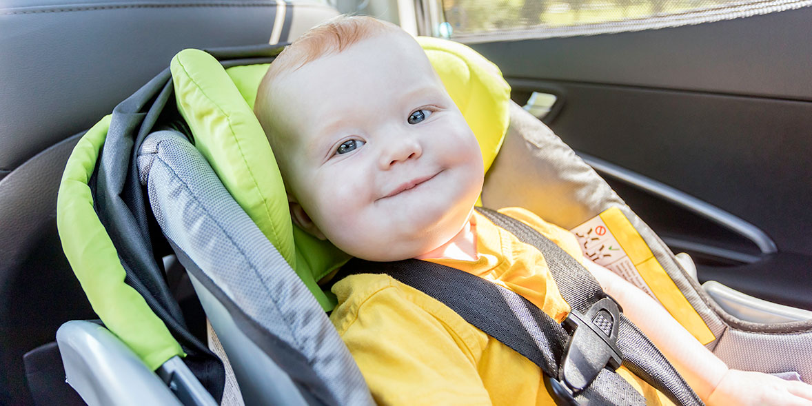 Smiling baby in a bright yellow shirt seated in a green and grey car seat, with sunlight streaming through a window.