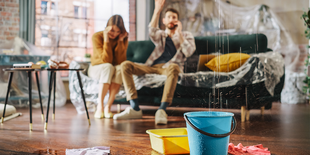 Water drips into a blue bucket on the floor, while a concerned woman sits beside a distressed man on a couch.