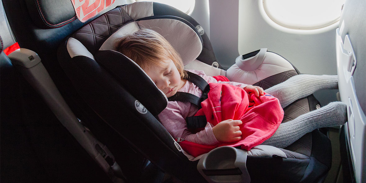 A young child sleeps in a car seat on an airplane, resting against the window with a red blanket draped over them.