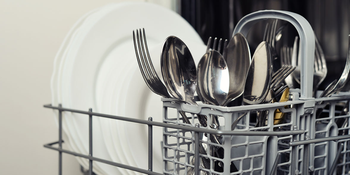 Silverware and plates arranged in a grey dishwasher basket, with forks, spoons, and knives visible alongside white dishes.