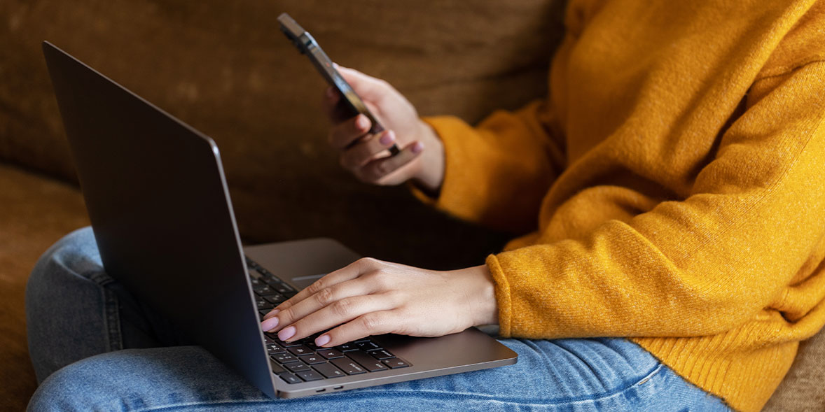 A person in a mustard sweater holds a smartphone while typing on a laptop, seated on a couch.