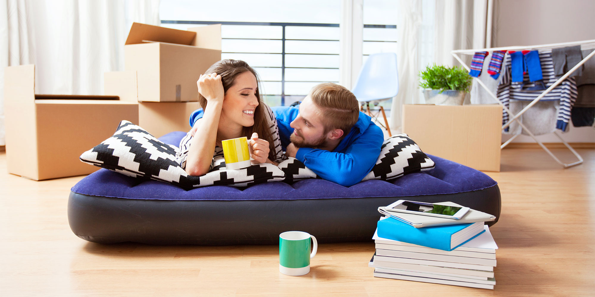 A couple relaxes on an inflatable bed, holding mugs, surrounded by moving boxes and laundry hanging nearby.