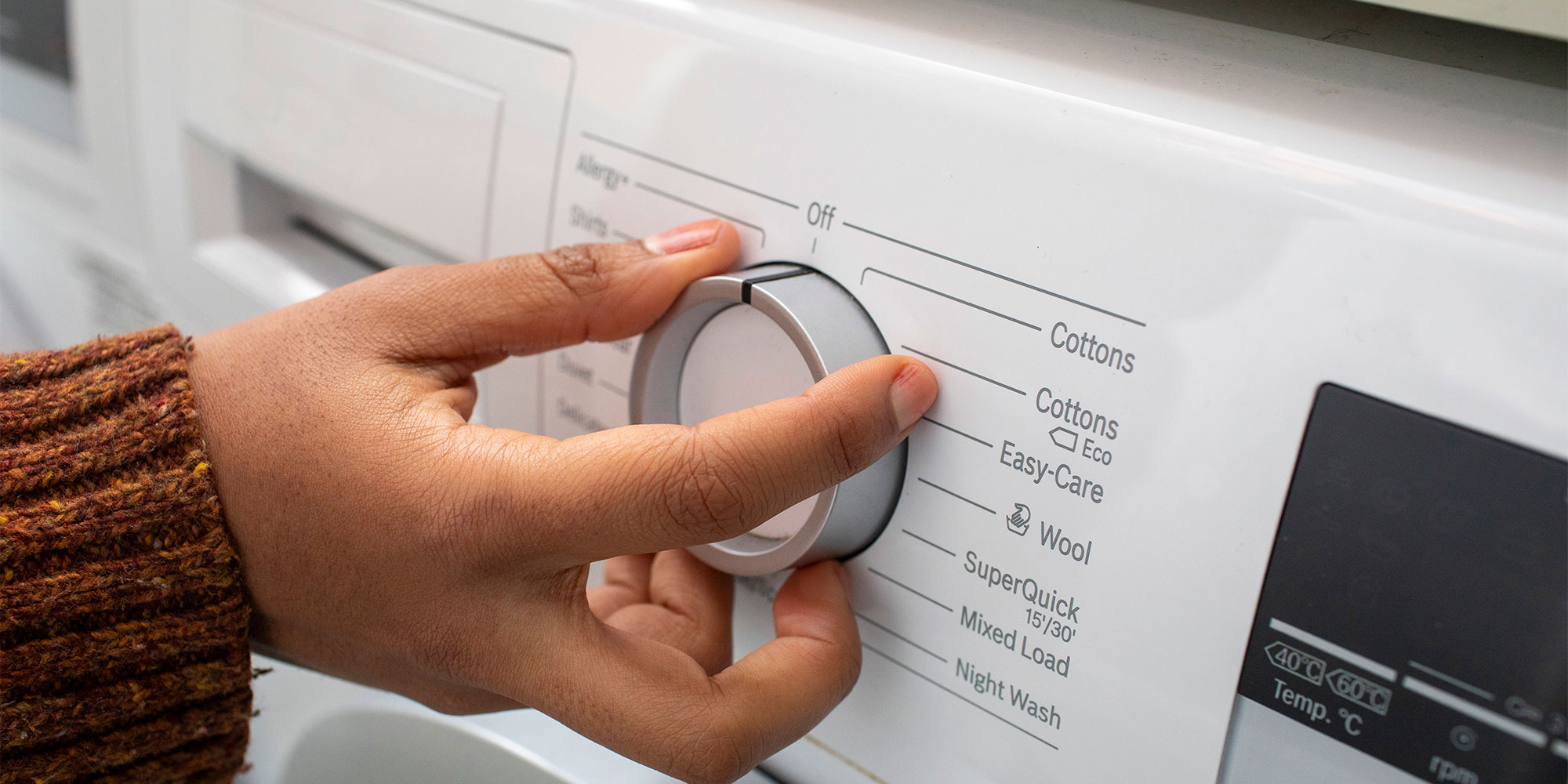 A hand adjusts the temperature dial on a washing machine, surrounded by icons representing different fabric types and cycles.