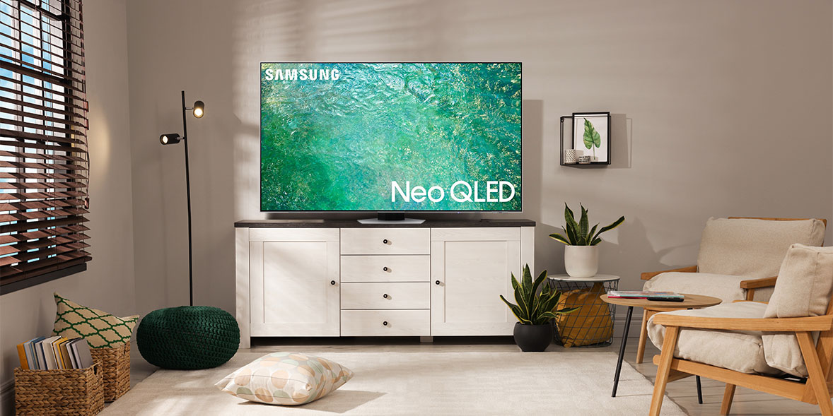 Samsung Neo QLED TV displayed on a white media console in a cozy living room with plants, a chair, and decorative lighting.