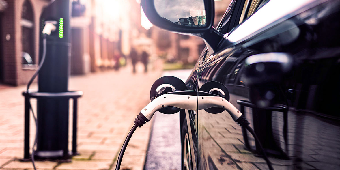 An electric car is plugged into a charging station on a city street, with a blurred background of pedestrians and buildings.