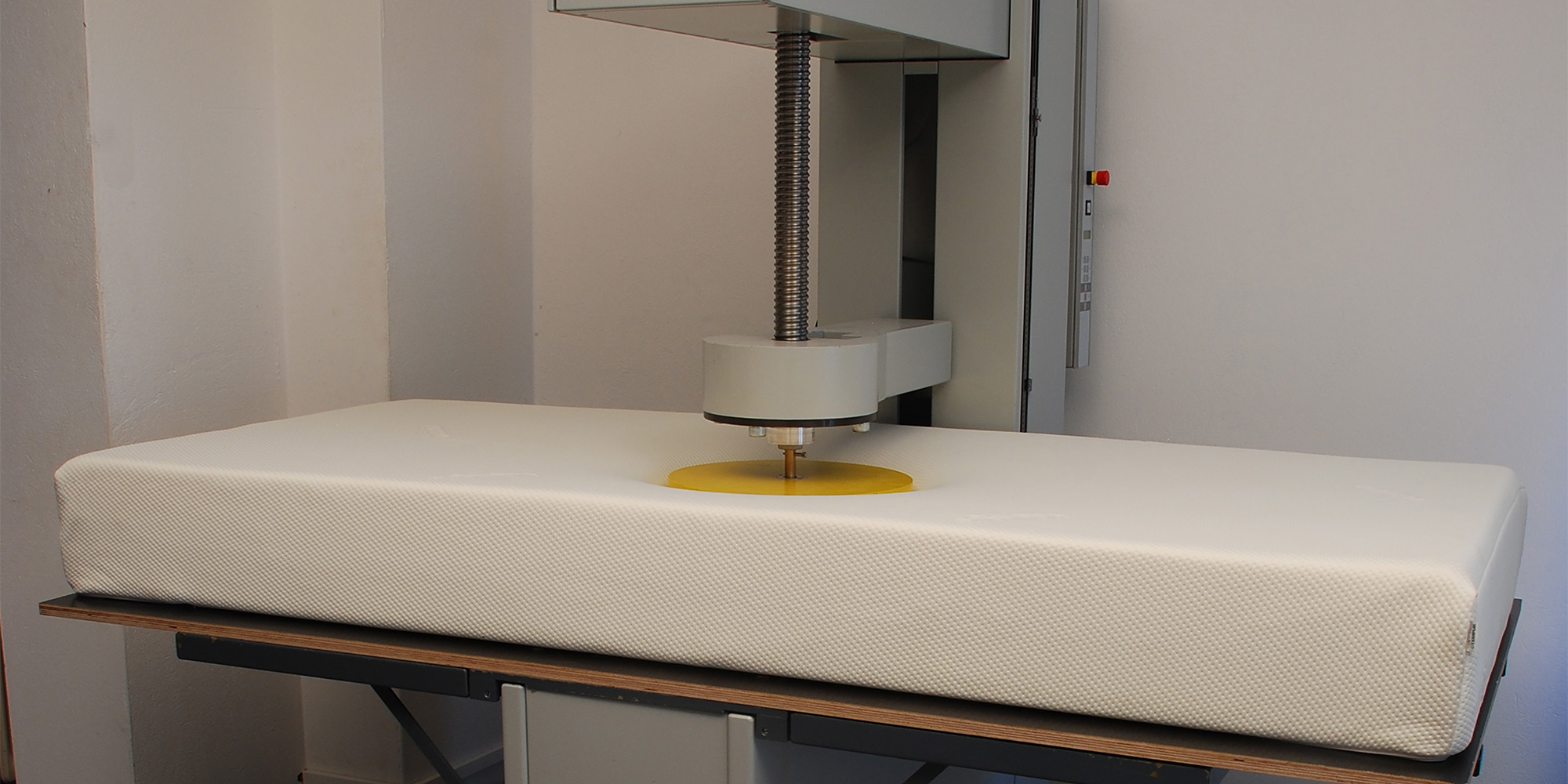 Compression testing machine applying pressure to a white mattress on a wooden table in a light-colored room.