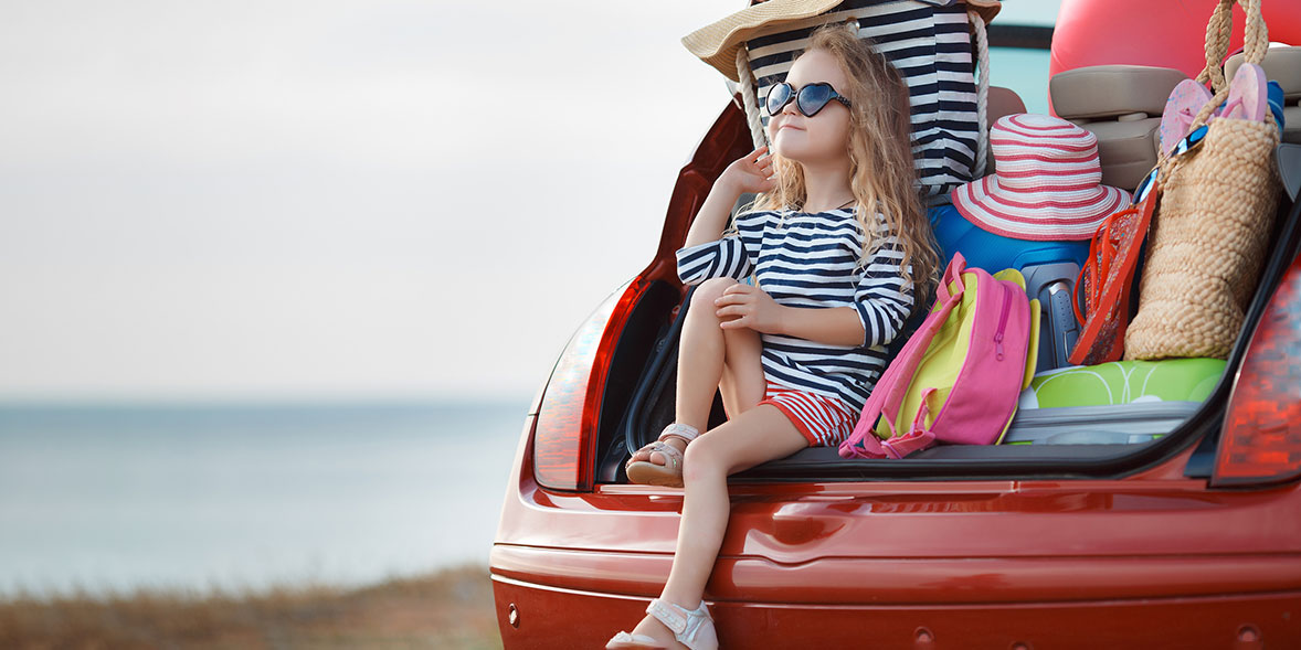 A young girl sits on the trunk of a car, surrounded by colorful beach bags and hats, with the ocean in the background.