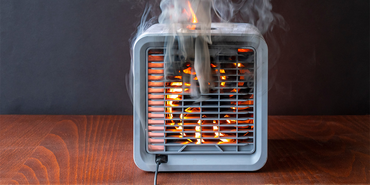 A small square heater with smoke and flames emerging from the front, set against a dark background on a wooden surface.