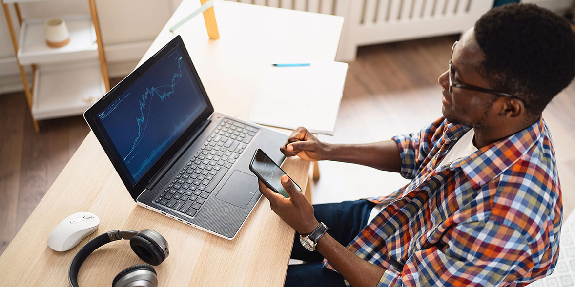 Man in a plaid shirt sitting at a desk, looking at a laptop displaying stock charts while holding a smartphone.