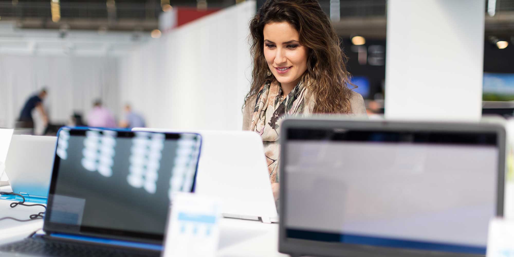 A man in a grey hoodie inspects laptops arranged on a table in a retail store, surrounded by tech accessories on display.