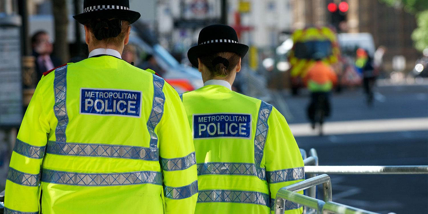 Two police officers in high-visibility yellow jackets with "METROPOLITAN POLICE" on the back, standing on a busy street.