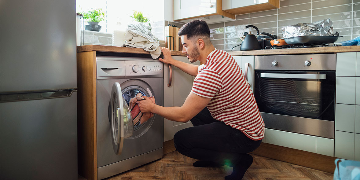 A man in a striped shirt crouches by a washing machine, loading laundry while a modern kitchen is visible in the background.