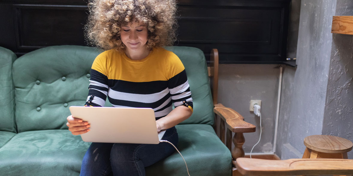 Woman with curly hair in a yellow and black striped shirt sits on a green couch, working on a laptop in a cozy setting.