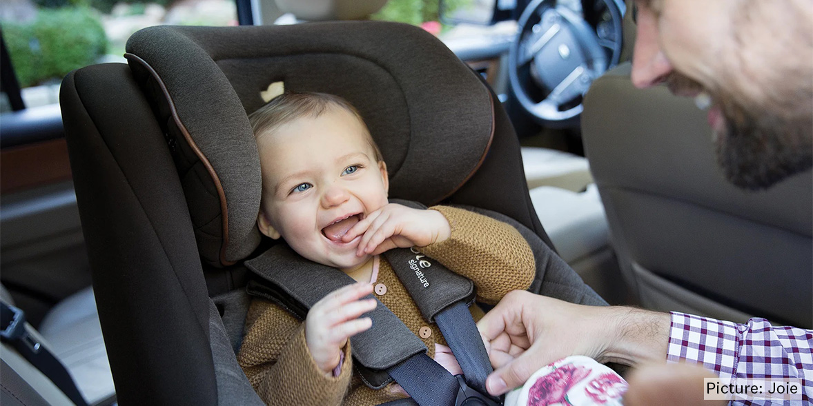 A joyful toddler in a car seat smiles and laughs while a caregiver playfully interacts with them.