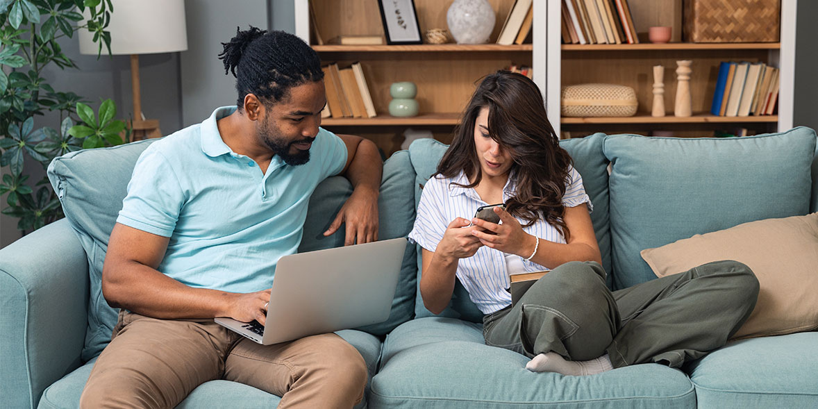 A man working on a laptop smiles at a woman sitting beside him, who is focused on her smartphone, both relaxed on a couch.
