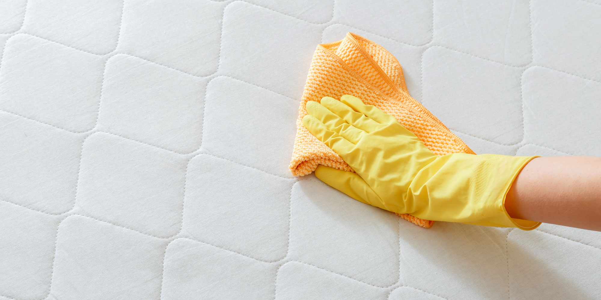 A hand wearing a yellow rubber glove wipes a textured white mattress with an orange cloth.