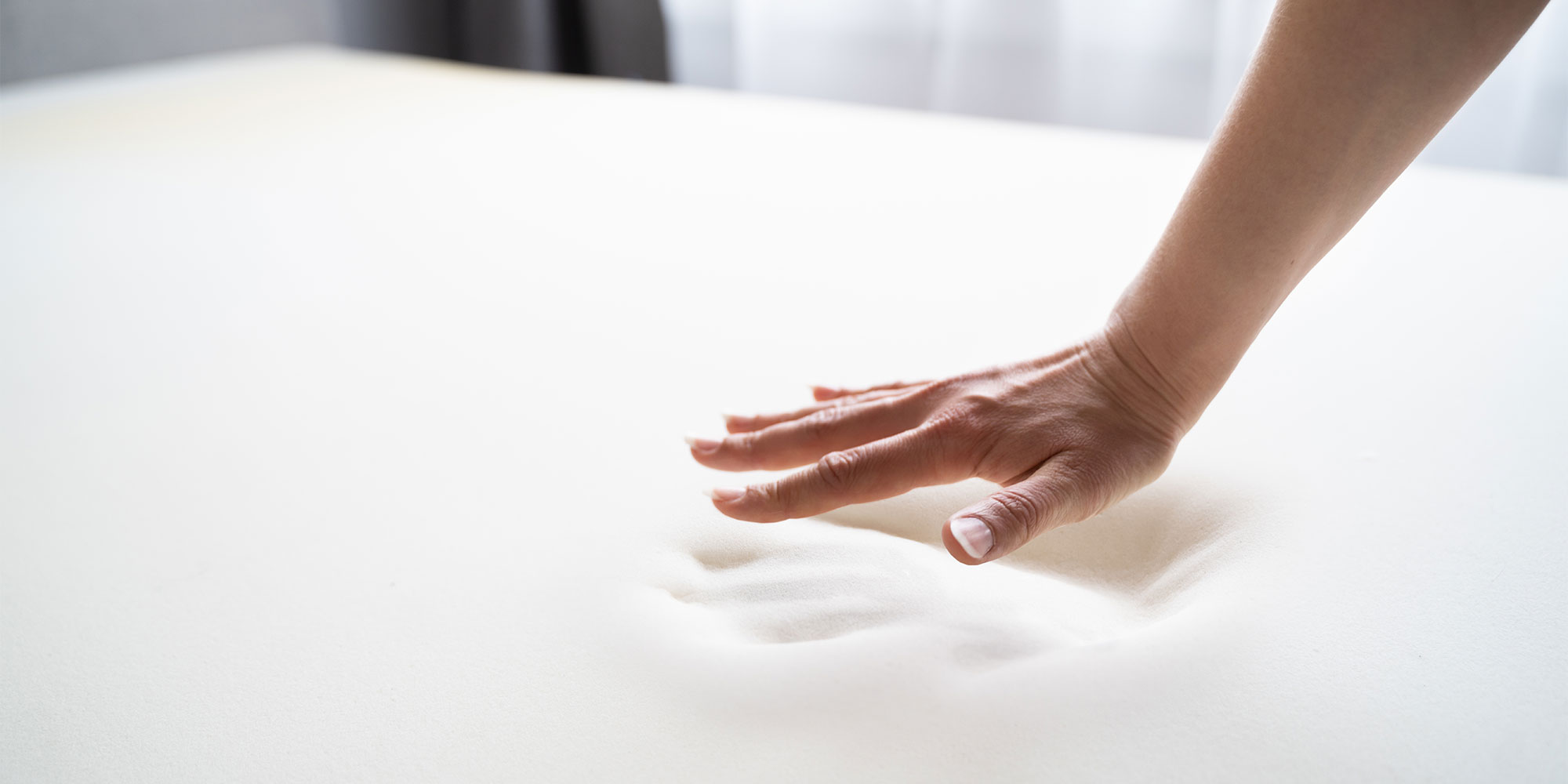 A hand presses into a soft, white memory foam mattress surface, leaving an imprint
