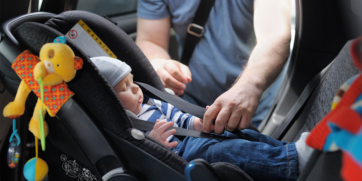Infant in a car seat, wearing a blue hat and striped shirt, as an adult's hand fastens the safety strap.