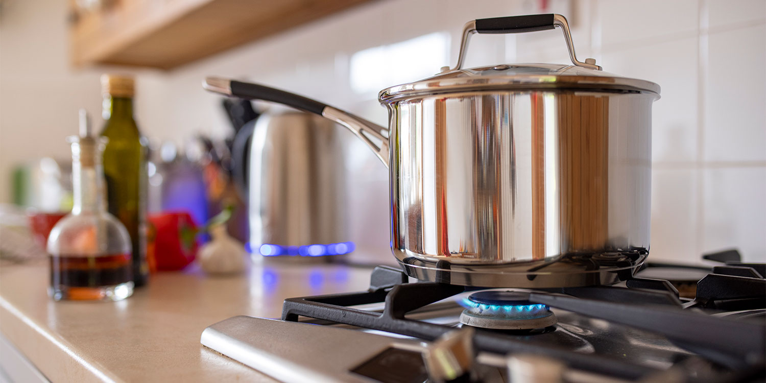 A stainless steel pot sits on a gas stove, with blue flames lit beneath it, surrounded by cooking oils and utensils.