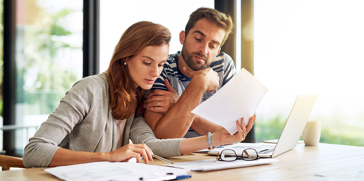 A woman and a man review documents together at a table, with a laptop and glasses nearby, in a sunlit room.