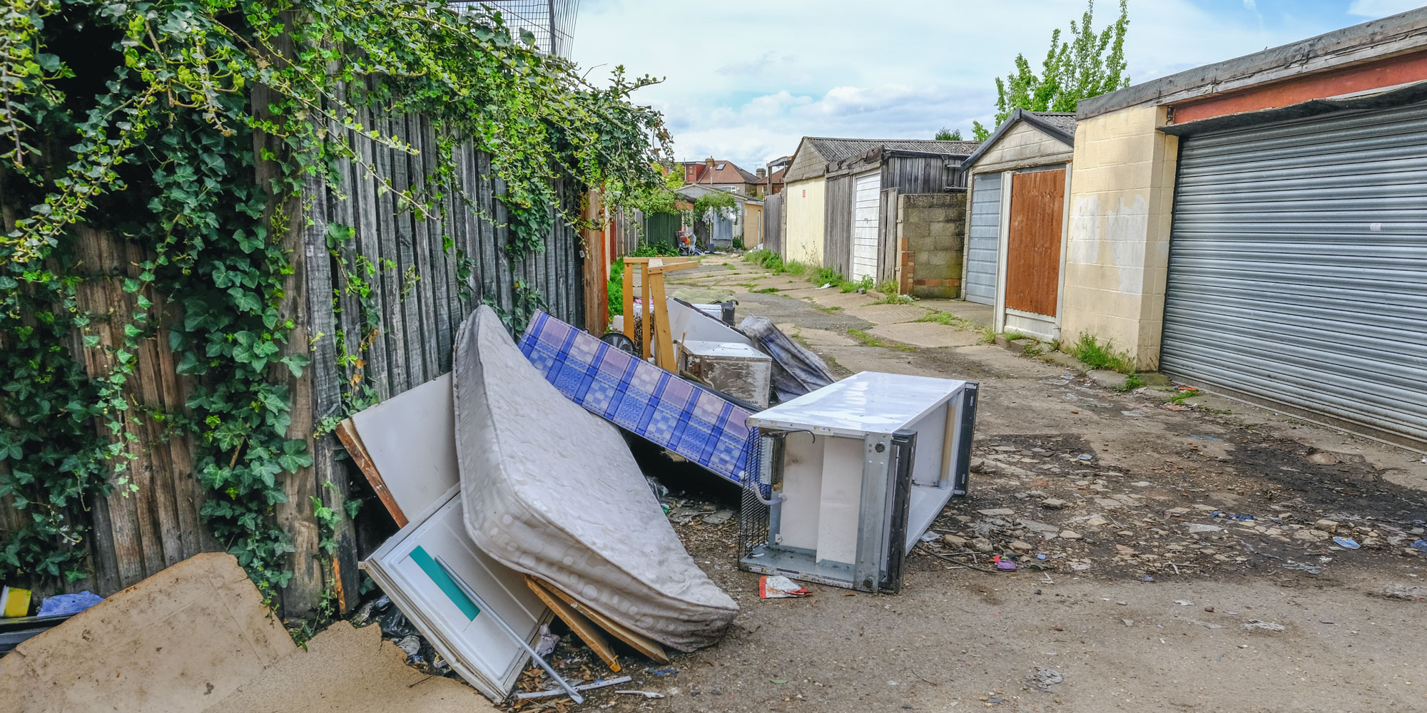 Discarded furniture, including a mattress and refrigerator, scattered in a narrow alleyway lined with garages and greenery.